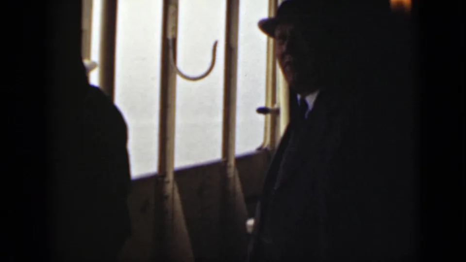 Adult man in a fedora and dark suit stands near bright vessel windows during the Ontario 1940 home-movie scene.