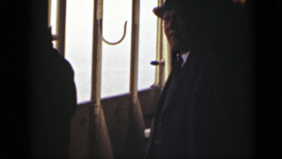 A passenger in dark clothing and a fedora is filmed close to the vessel windows, with strong backlight and soft 8mm grain.