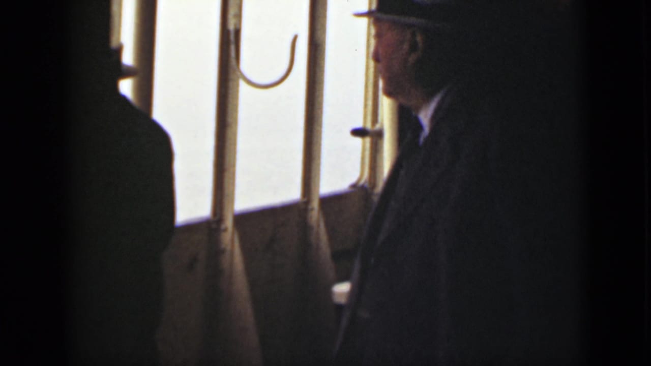 Suited man in a fedora standing beside a bright boat window.