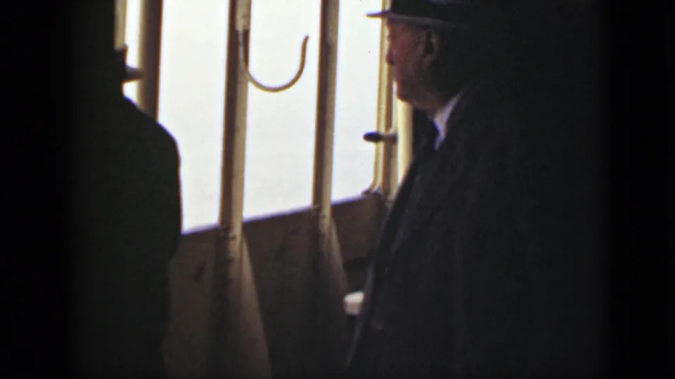 In 1940 Ontario home-movie footage, a man in a dark suit and fedora stands near a boat window with bright water outside.