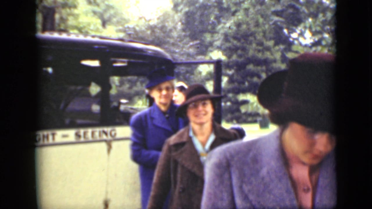 Women in period coats and hats stand near a sightseeing bus in a park-like setting.