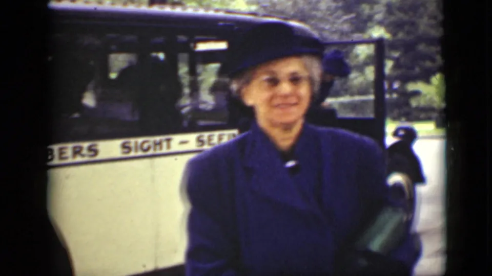 1940 Ontario frame of a smiling woman in a blue coat and hat near a cream sightseeing bus.