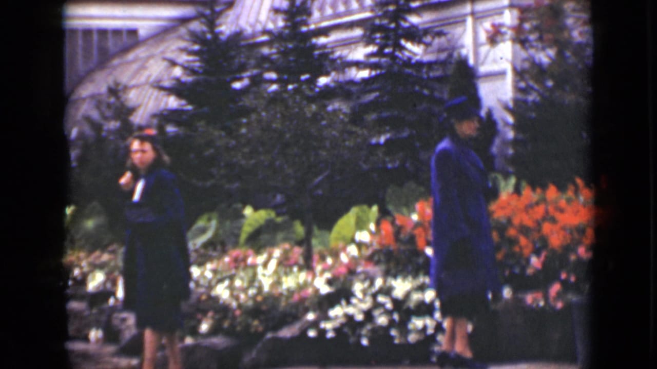 Two women in blue outerwear stand and walk near flower beds and evergreen plantings.