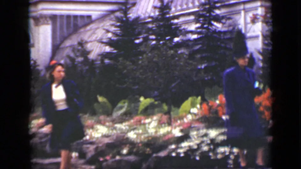 A woman in a blue coat and white blouse walks through a flower garden, while another blue-clad figure is partly visible near the shrubs.