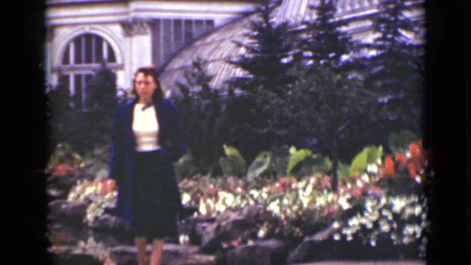 A woman in a blue coat, white blouse, and dark skirt stands near flower beds in an Ontario garden, with shrubs and an arched conservatory window behind her.