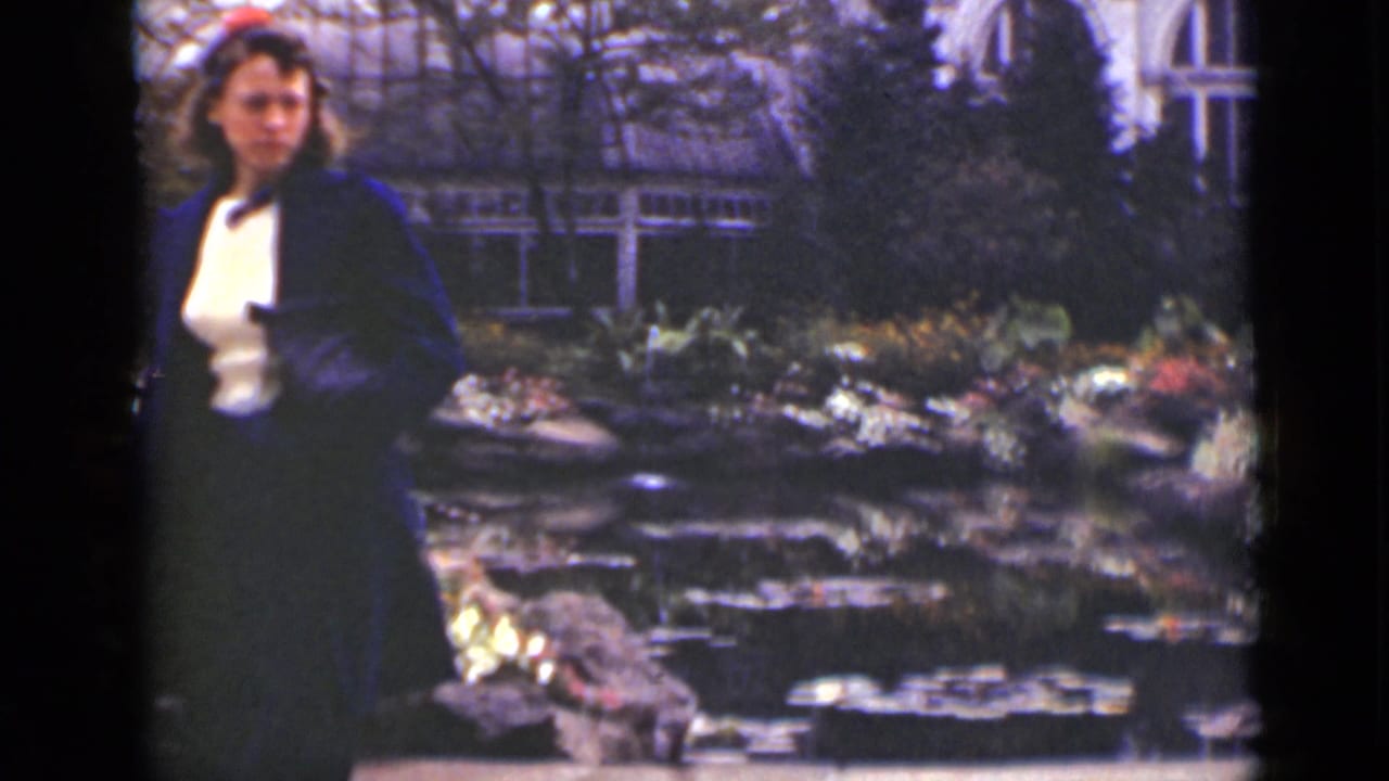 Woman in a dark coat stands beside a garden pond with greenhouse windows behind her.