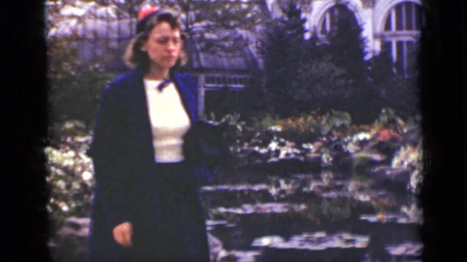 A woman in period dress walks through an Ontario garden, passing pond stones and greenhouse windows.
