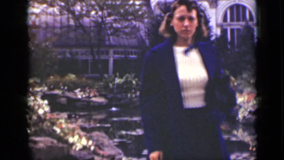 Closer 1940s Ontario view of the woman facing the camera near rocks, pond plantings, and greenhouse glazing.