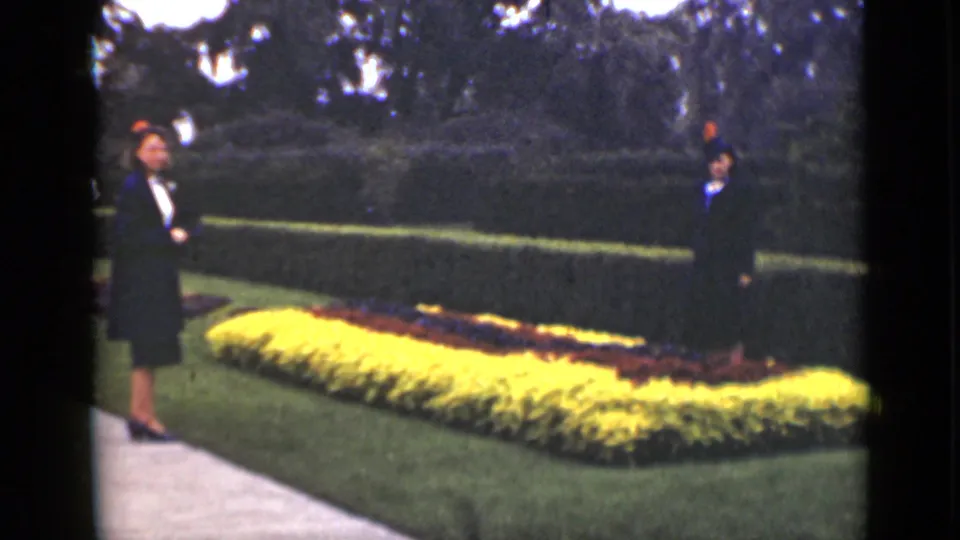 Two adults stand in a formal Ontario garden near clipped hedges, lawn, path, and a bright yellow flower bed.