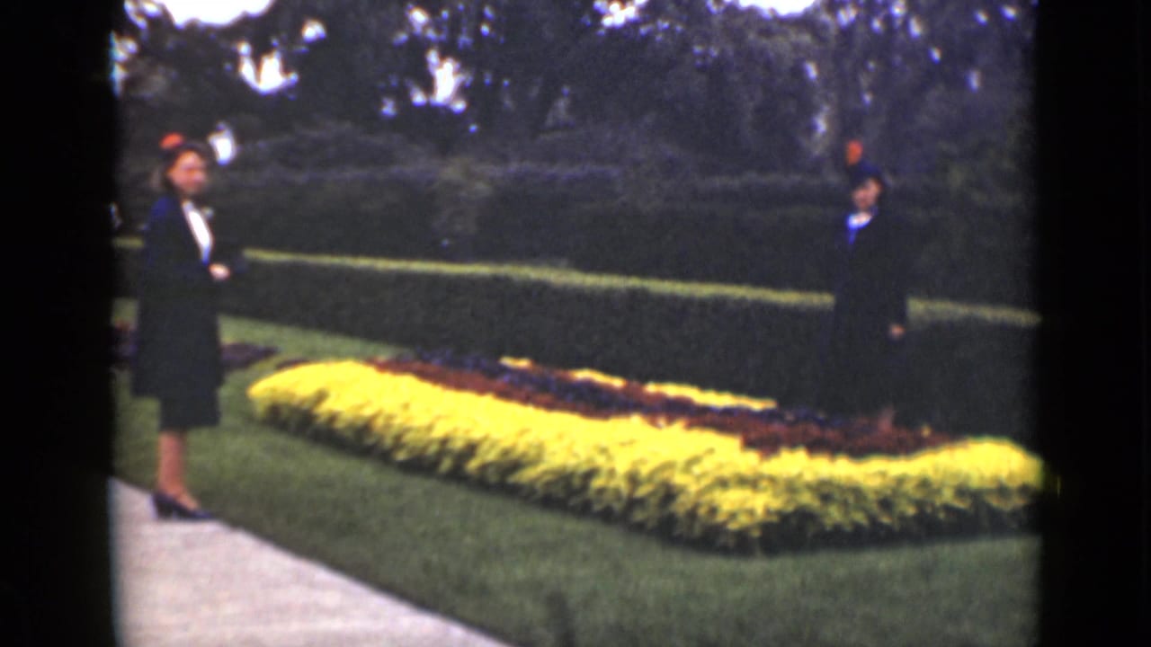 Two women in dark dresses and hats stand beside a bright flower bed and hedge.