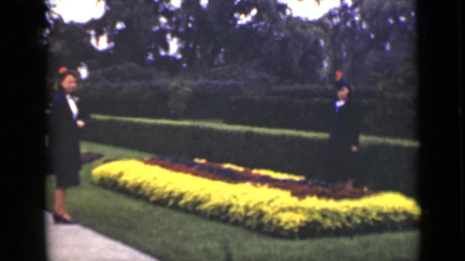 Two formally dressed women appear in an Ontario garden, framed by a clipped hedge, lawn, paved walk, and vivid flower beds.