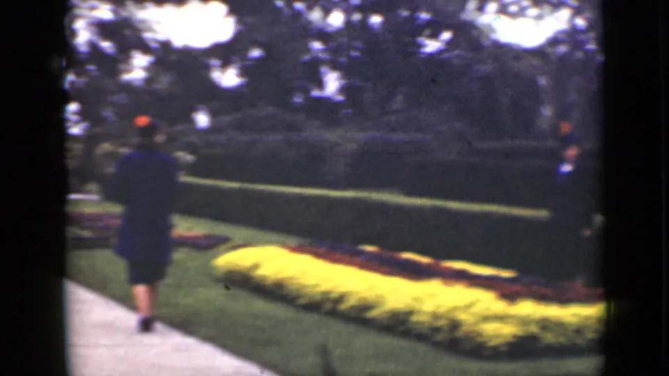 One woman in dark formal clothing turns away on the garden path as another remains near the hedge and flower beds.