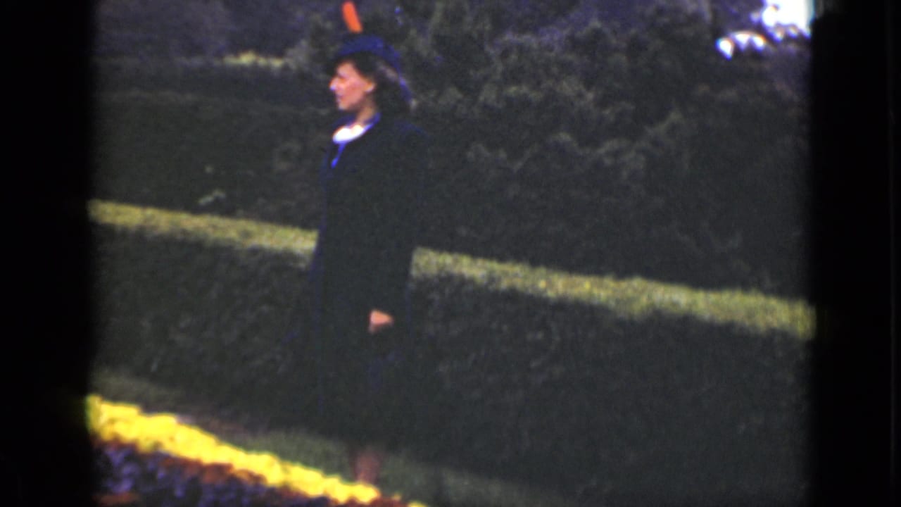 Woman in dark coat and hat seen in profile beside a hedge and low flowerbed.