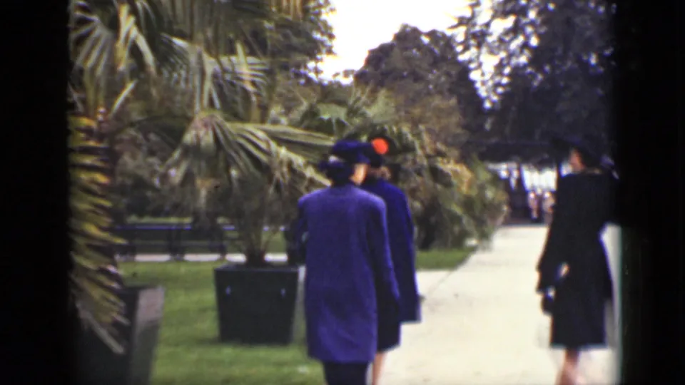 Women in formal coats walk along a garden path in Ontario circa 1940, with palms, hedges, lawns, and other visitors visible ahead.
