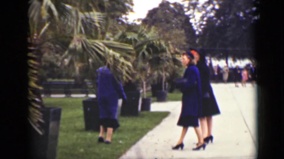 1940 Ontario 8mm frame of women in coats walking through a landscaped square with planters and palm-like foliage.
