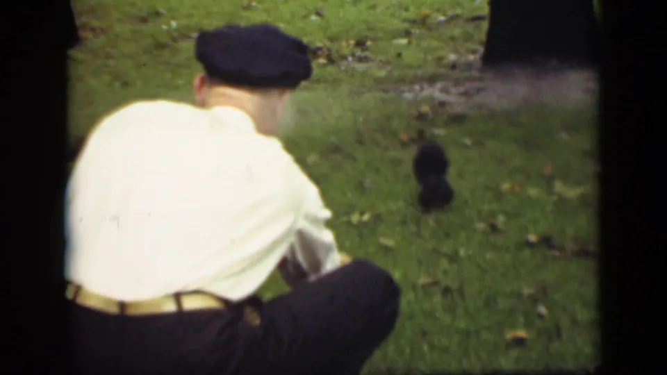 A man in a cap crouches on the grass in the same 1940 Ontario home-movie clip, looking toward a small dark object.