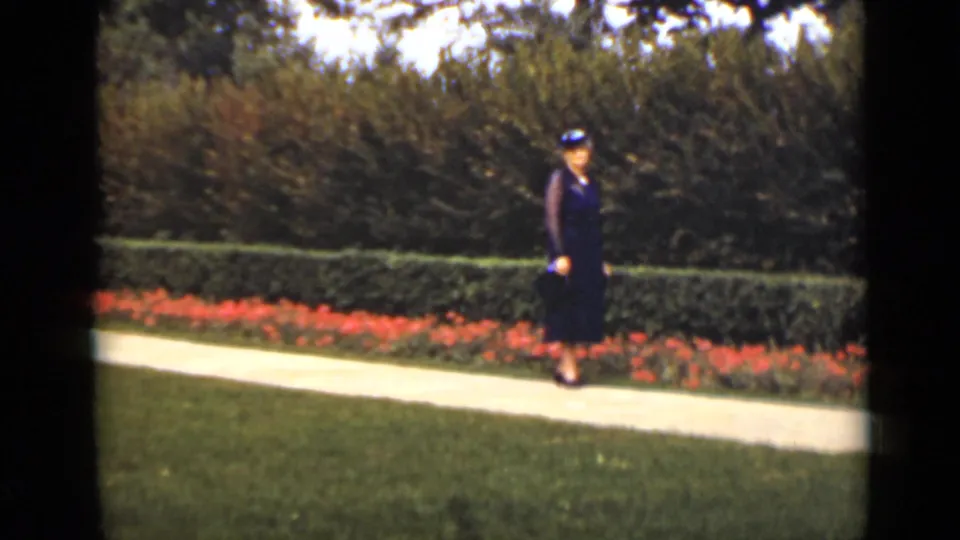 The woman walks in a manicured garden, with red flowers and green hedges visible in this 1940s Ontario home movie.