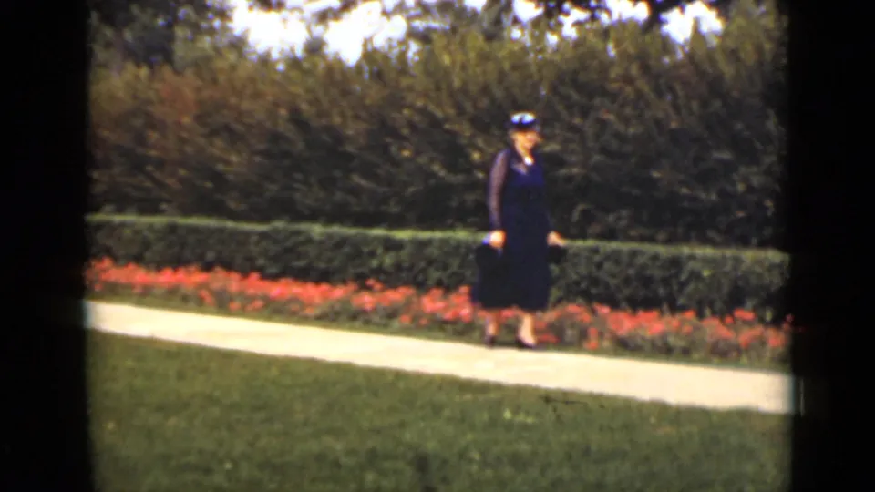 A woman continues walking along a paved garden path, framed by lawn, red flowers, and hedges in 1940s Ontario footage.