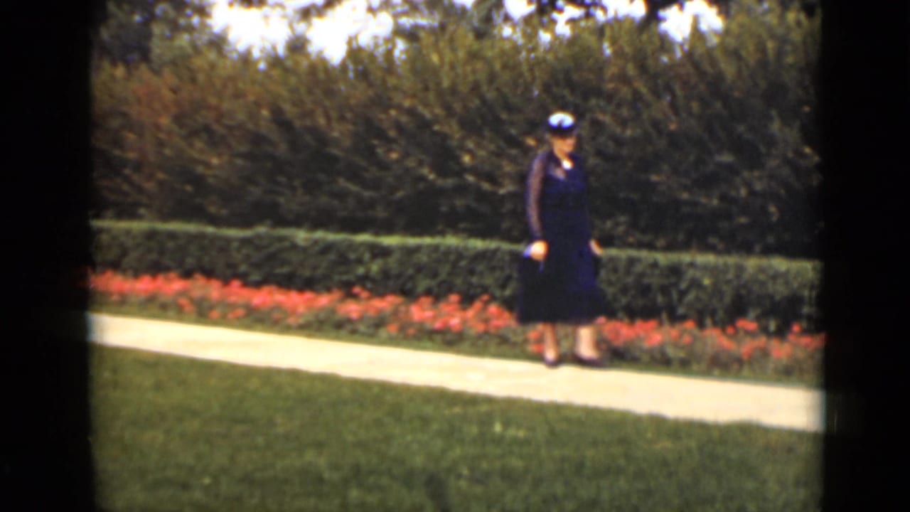 Woman in a dark dress and hat walking beside red flowers and hedges.