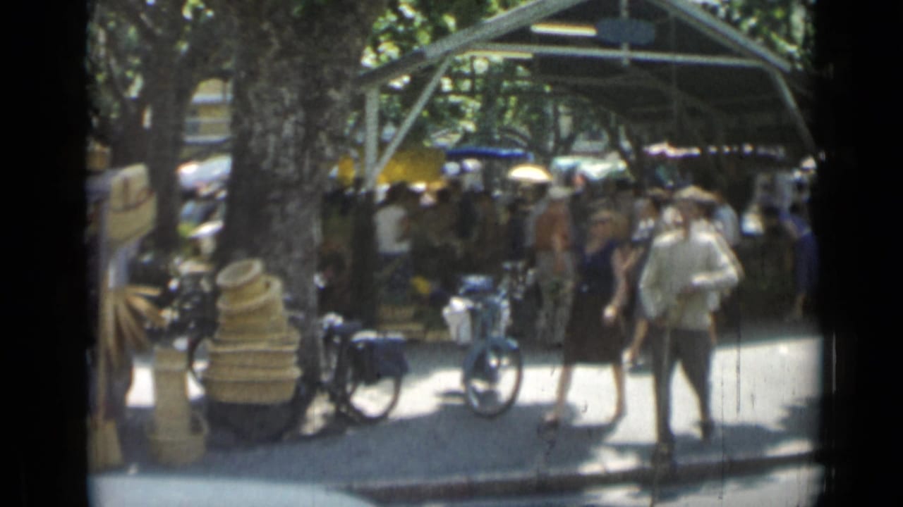 People walk past a shaded market stall with baskets and bicycles nearby.