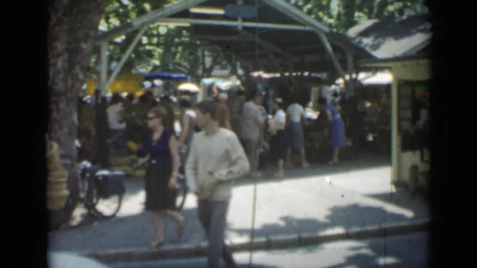 A man and woman walk past a crowded 1962 Cannes open-air market with bicycles, baskets, trees, and stalls behind them.