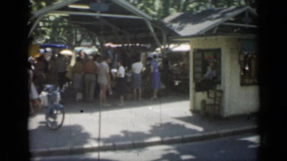 Open-air Cannes market view with shoppers under canopies, a small kiosk, bicycles, trees, and bright pavement.