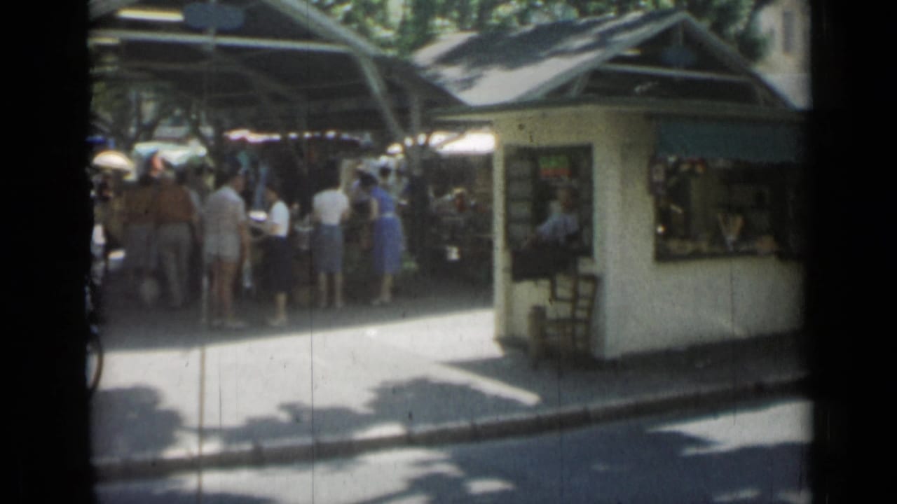 White kiosk with green awning beside a shaded outdoor market and pedestrians.