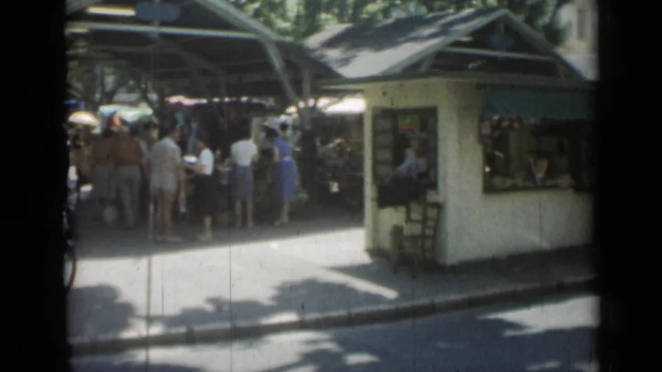 1962 Cannes street view with a white kiosk, green awning, market crowd, trees, and bright pavement.