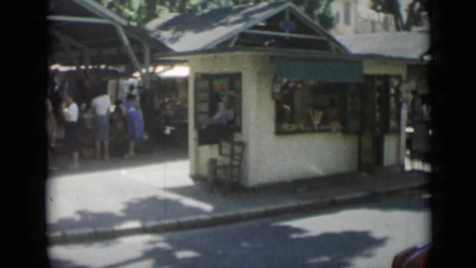 Cannes street-market frame showing a kiosk, shaded stalls, pedestrians, and strong sun on the pavement.