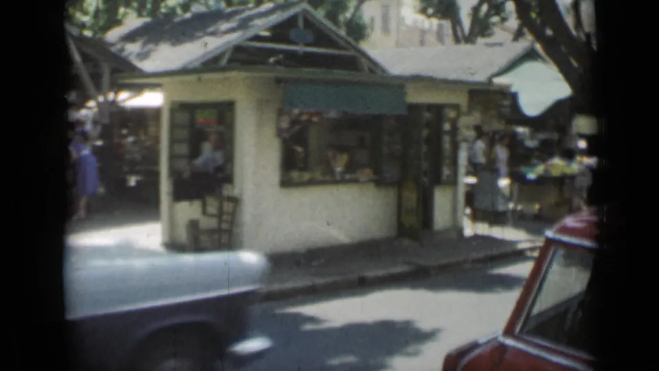 1962 Cannes market street with a kiosk, pedestrians, produce or goods tables, and cars moving through the foreground.