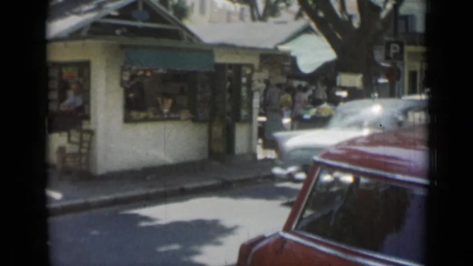 Cannes, 1962, with a red car passing a white kiosk near market stalls, pedestrians, trees, and a parking sign.
