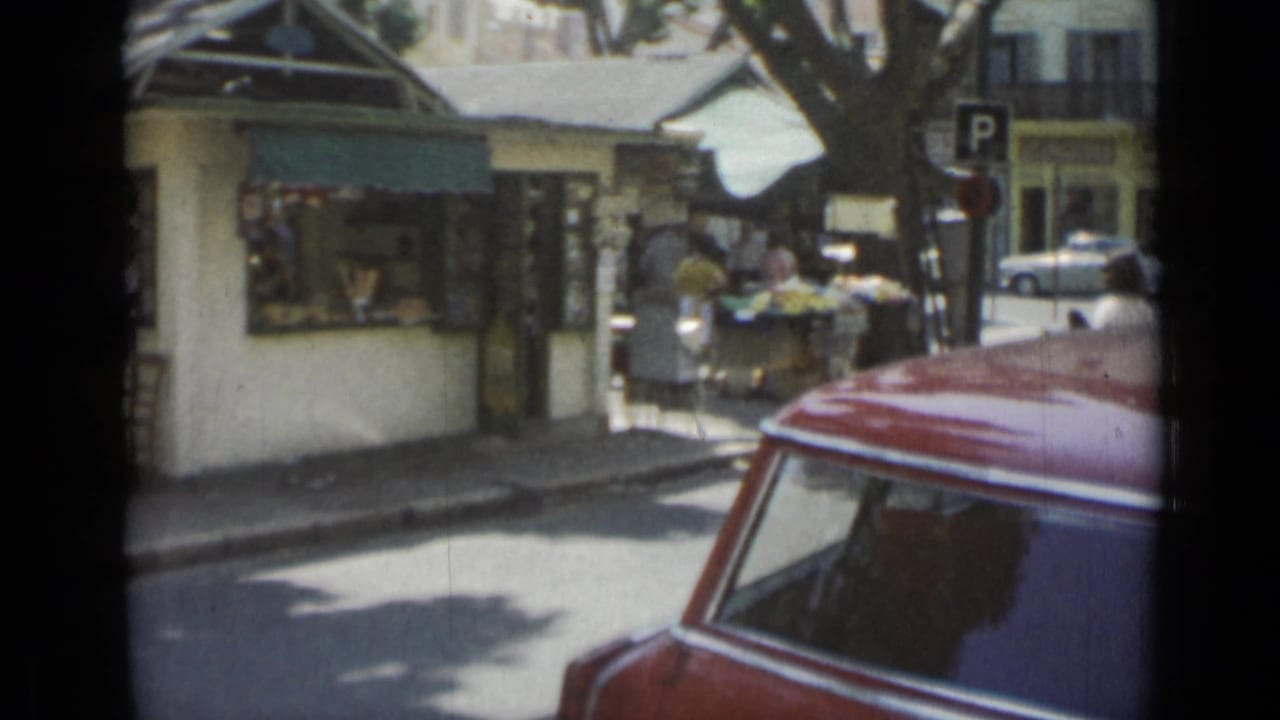 Street corner with market stalls, pedestrians, a parking sign, and a red car roof in the foreground.