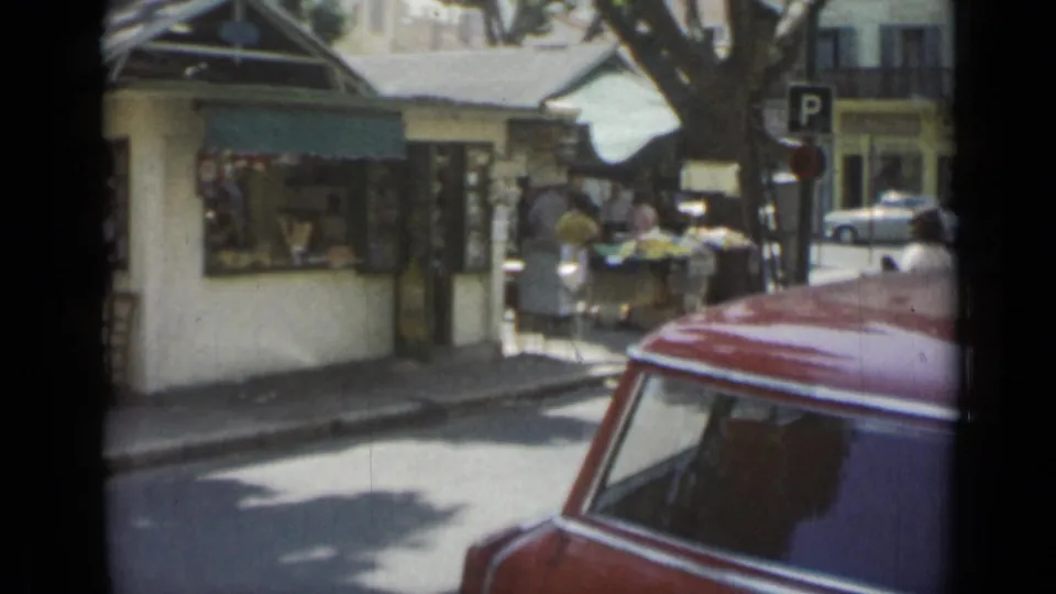 1962 Cannes street view with pedestrians near market stalls, a parking sign, storefronts, trees, and a red car passing close to the camera.