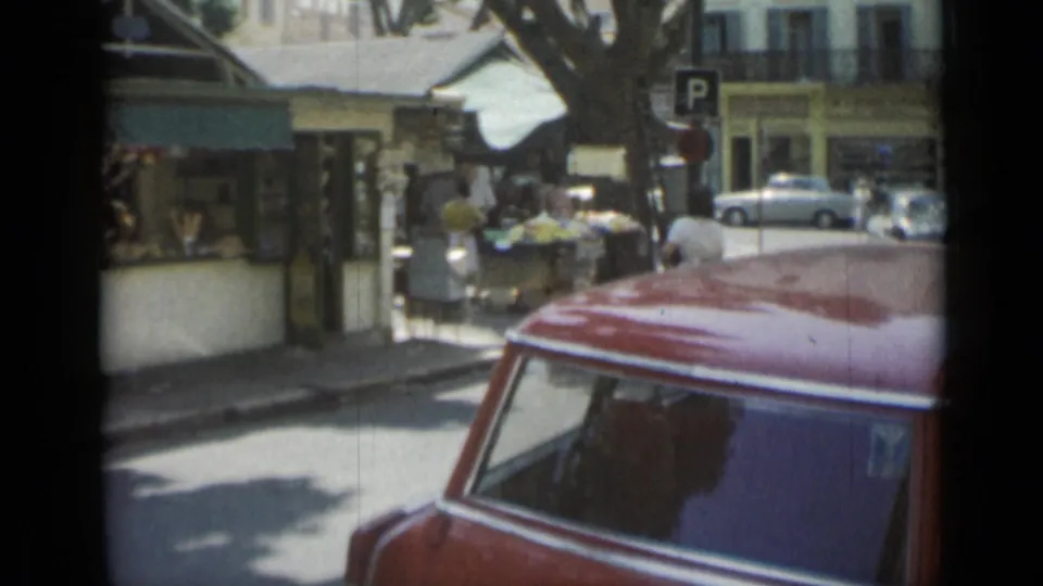 Cannes street scene showing people near market stalls and storefronts, with parked cars, shade trees, and a red vehicle partly blocking the lower frame.