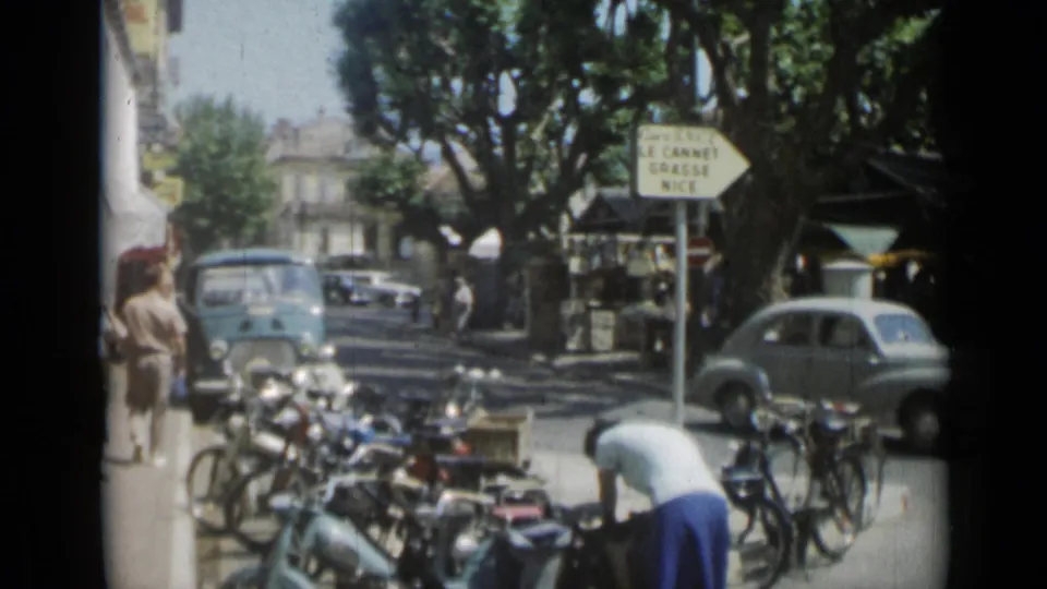 1962 Cannes street corner with parked bicycles, compact cars, pedestrians, a road sign toward Le Cannet, Grasse, and Nice, and shaded stalls.