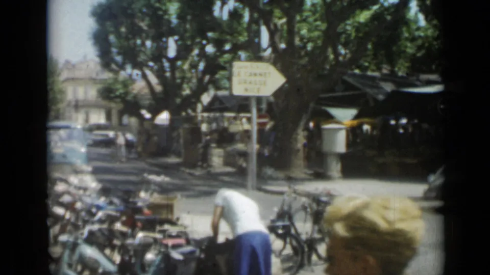 Cannes street view with bicycles in the foreground, a person bending near the curb, compact cars, market stalls, trees, and a road sign for nearby towns.