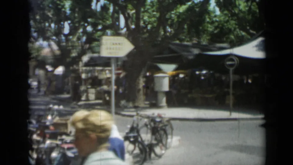 1962 Cannes street scene with market tents, parked bicycles, pedestrians, and French directional signage under leafy trees.