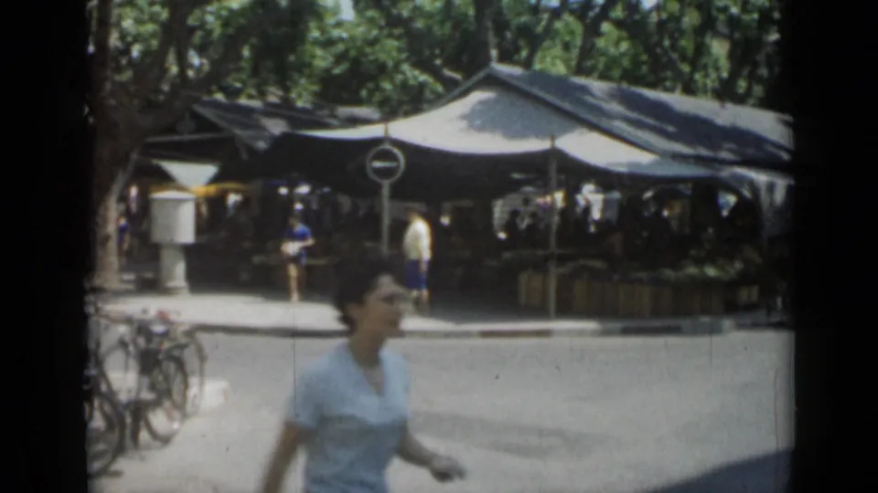 Cannes street market view with a woman in the foreground, bicycles at the curb, and shaded tents beneath trees in 1962.