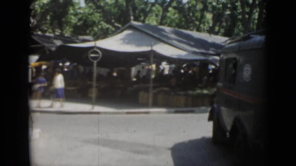 Dark vehicle passing a shaded Cannes market with pedestrians, road signs, trees, and street shadows in 1962 home-movie footage.