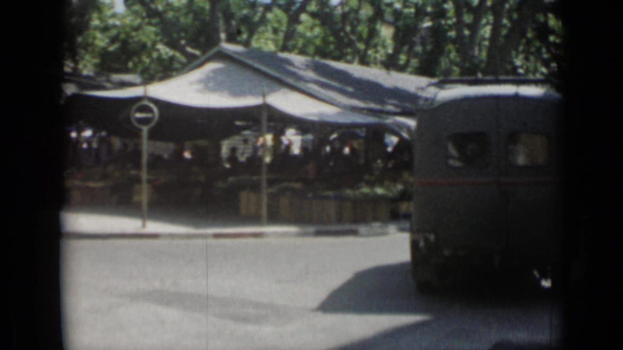 1962 Cannes street view with a dark van turning past canvas market tents and pedestrians under trees.