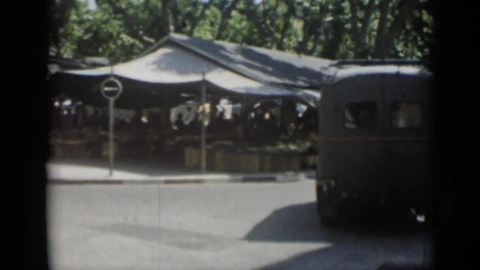 1962 Cannes street view with a dark van turning past canvas market tents and pedestrians under trees.