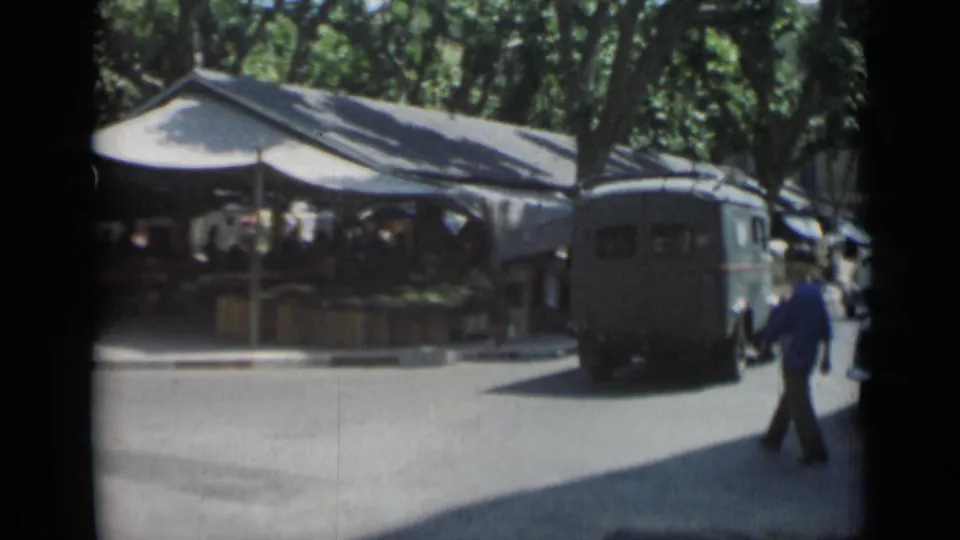 1962 Cannes market street with a dark van, a man in a blue shirt, and tents along the curb.