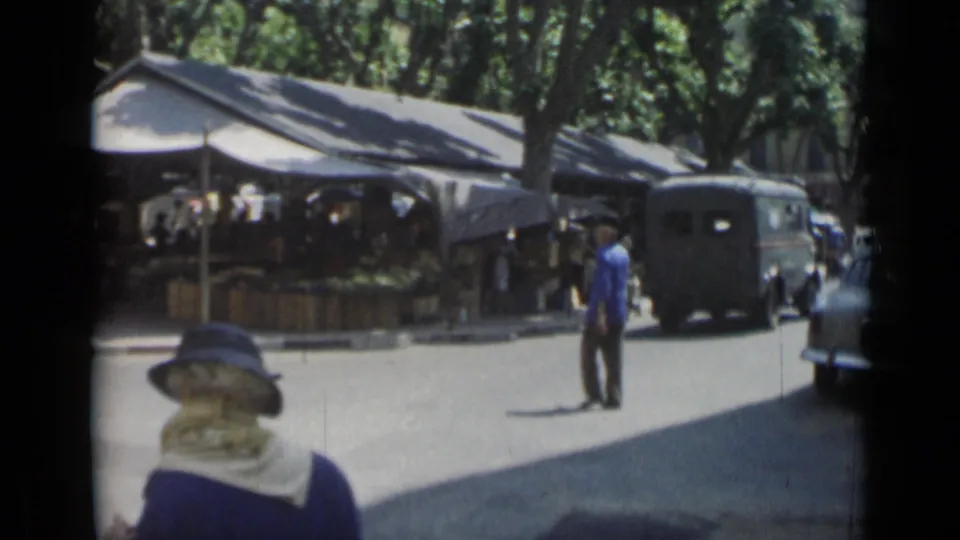 1962 Cannes street scene with a woman in a hat, a passing van, parked cars, and shaded market tents.