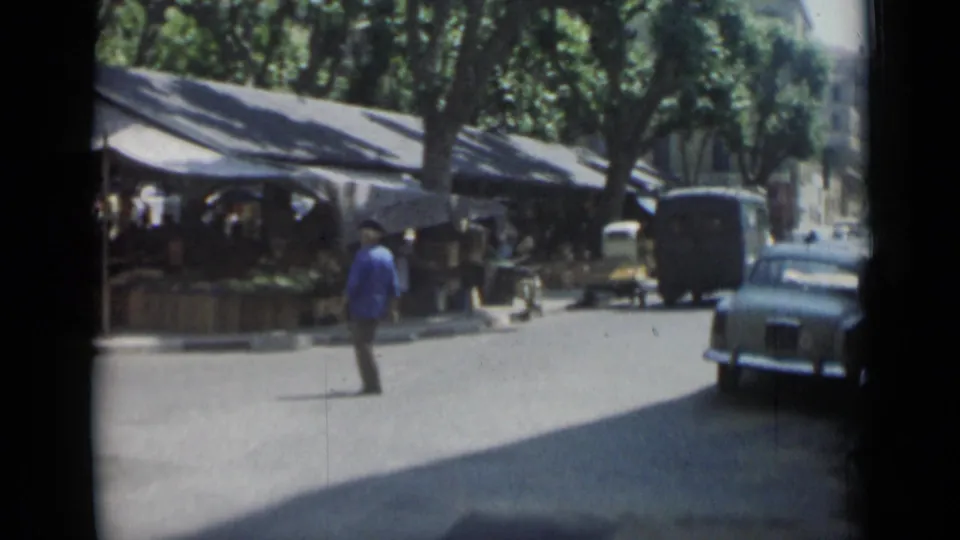 1962 Cannes market street with pedestrians, parked cars, a small yellow vehicle, and a van farther along the curb.