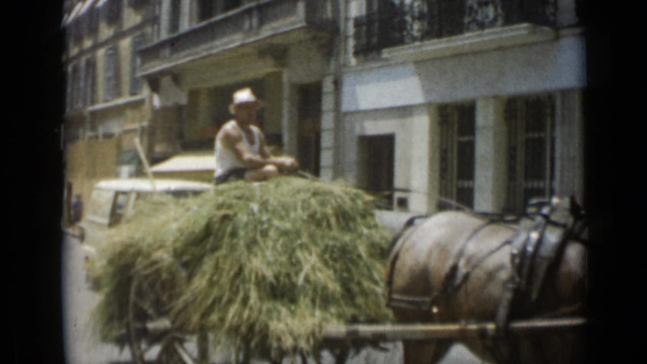 Man riding a hay-loaded horse-drawn cart along a city street.