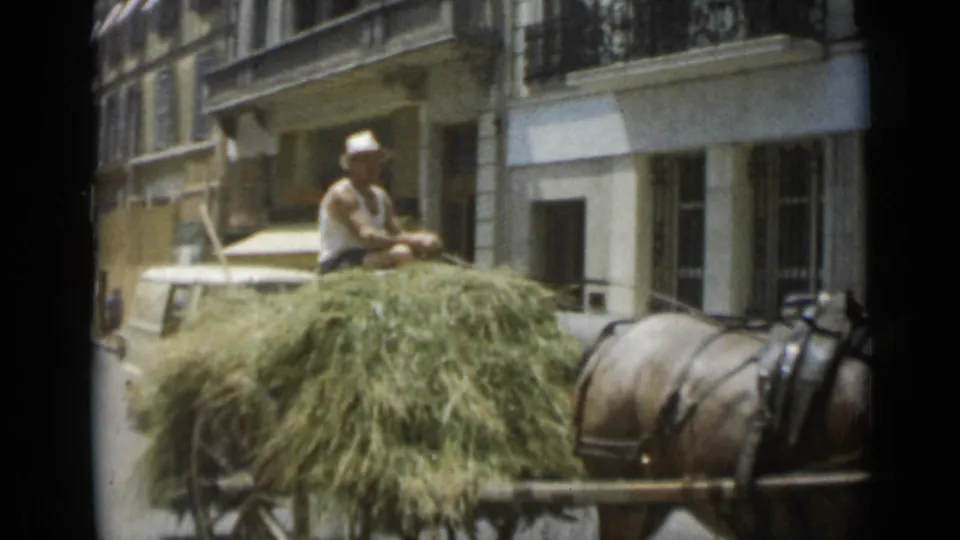 In Cannes, France in 1962, a man in a hat rides a hay-filled horse-drawn cart past multi-story street facades.