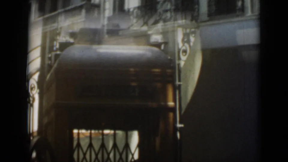 A warm, dim 1962 interior view shows a vintage elevator with a diamond-pattern metal gate and decorative side details.