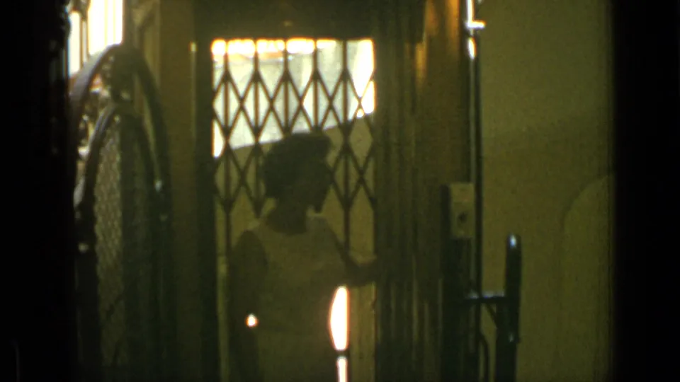 A silhouetted woman stands behind the diamond-pattern gate of a vintage elevator, lit by warm interior light in 1962 Cannes footage.
