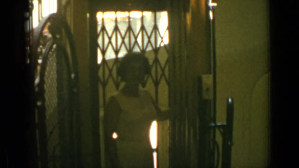 In Cannes 1962 footage, a woman in a light dress stands inside a doorway behind a diamond-patterned metal gate.