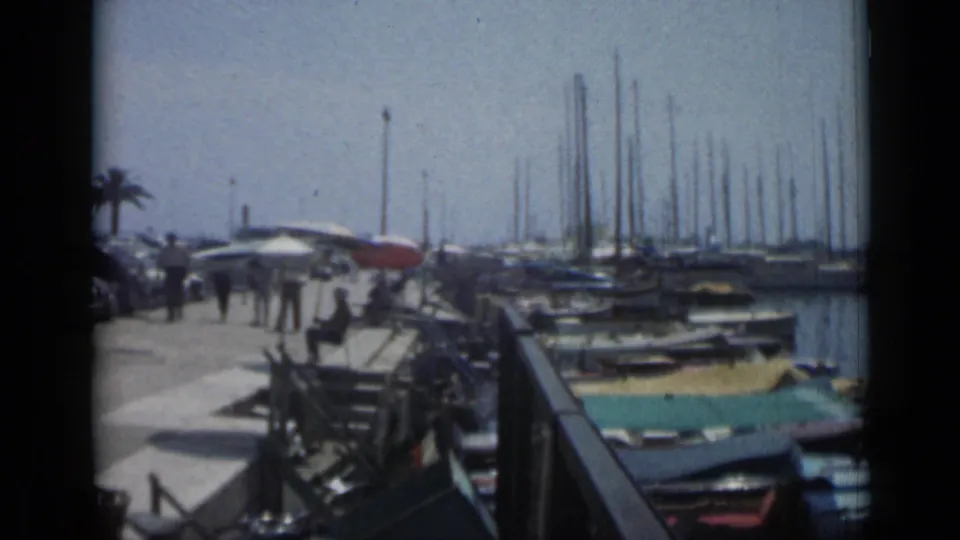 A Cannes marina view shows docked small boats, many sailboat masts, pedestrians on the pier, and calm water in faded 8mm color.